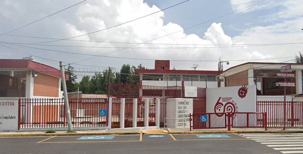 Fotografía en la que se muestra la fachada del plantel, conformada por muros de color blanco intercalados con rejas de color rojo. Una de las rejas da acceso al plantel, y el muro que está a su izquierda tiene el logo de la DGCFT.